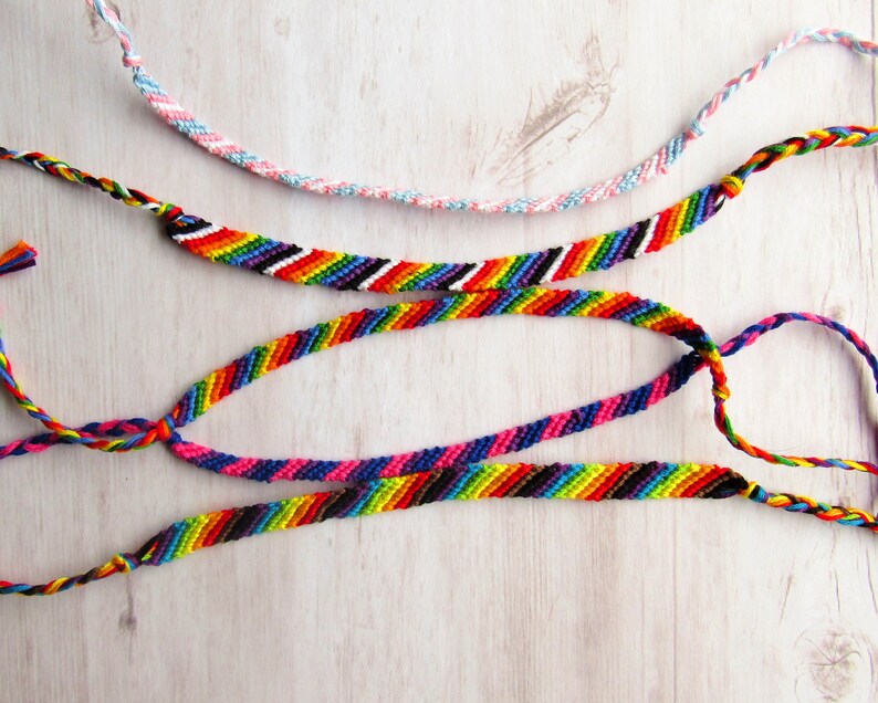 May include: Four handmade friendship bracelets made of woven embroidery floss. One bracelet features a light blue, pink, and white twisted pattern. Three bracelets display diagonal stripes in vibrant rainbow colors, including red, orange, yellow, green, blue, and purple.