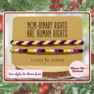 May include: Two non-binary pride bracelets made of braided thread in yellow, white, purple, and black. The bracelets are displayed on a rectangular kraft paper card with the text "NON-BINARY RIGHTS ARE HUMAN RIGHTS" and "Florence Mae Handmade".