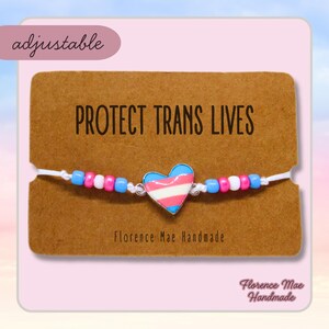 May include: A white string bracelet with blue and pink beads and a heart charm with the transgender pride flag colors. The bracelet is on a brown card with the text "PROTECT TRANS LIVES" and "Florence Mae Handmade".