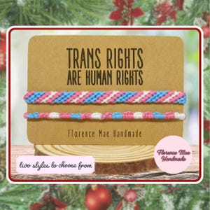 May include: Two woven bracelets featuring the trans pride flag colors of light blue, pink, and white. The bracelets are displayed on a rectangular brown card with the text "TRANS RIGHTS ARE HUMAN RIGHTS" and "Florence Mae Handmade" printed on it.