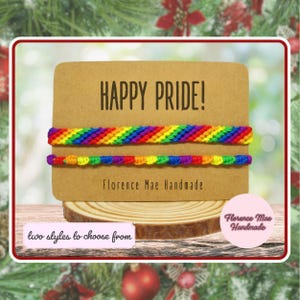 May include: Two rainbow-colored woven bracelets displayed on a rectangular tan card. The card features the text "HAPPY PRIDE!" and "Florence Mae Handmade". One bracelet has a diagonal striped pattern, while the other has a knotted texture.