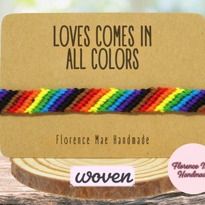 May include: A woven bracelet featuring a diagonal striped pattern in rainbow colors including red, orange, yellow, green, blue, purple, brown, and black. The bracelet is displayed on a rectangular tan card with the text LOVES COMES IN ALL COLORS.
