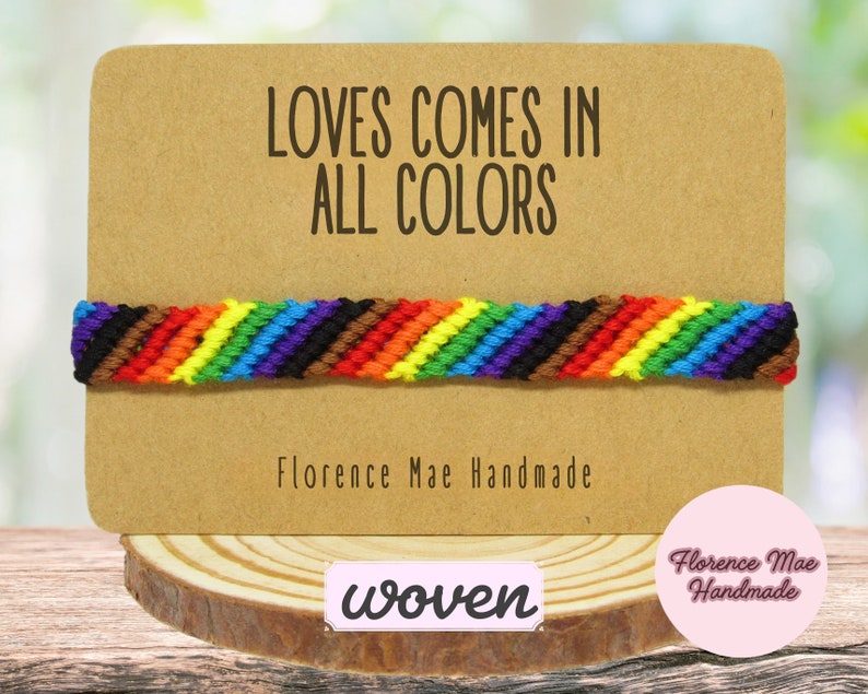 May include: A woven bracelet featuring a diagonal striped pattern in rainbow colors including red, orange, yellow, green, blue, purple, brown, and black. The bracelet is displayed on a rectangular tan card with the text LOVES COMES IN ALL COLORS.