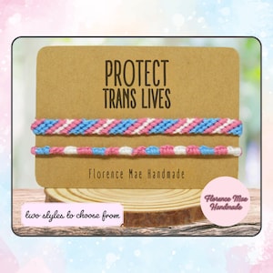May include: Two friendship bracelets with a blue, pink, and white color scheme. The bracelets are displayed on a brown card with the text "PROTECT TRANS LIVES" and "Florence Mae Handmade".