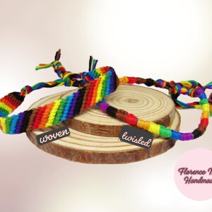 May include: Two rainbow-colored bracelets made of thread. One bracelet features a flat woven pattern with a black tag labeled "woven," while the other is a round twisted cord with a black tag labeled "twisted." A pink circle displays "Florence Mae Handmade."