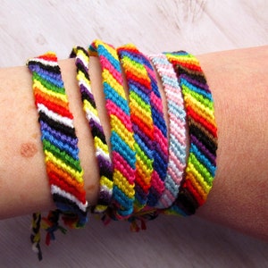 May include: A collection of six handmade friendship bracelets featuring various diagonal striped patterns in vibrant colors including red, orange, yellow, green, blue, purple, black, white, and pink, all crafted from woven embroidery thread.