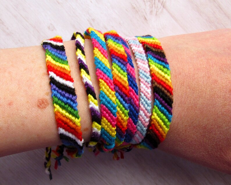 May include: A collection of six handmade friendship bracelets featuring various diagonal striped patterns in vibrant colors including red, orange, yellow, green, blue, purple, black, white, and pink, all crafted from woven embroidery thread.