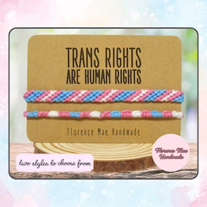 May include: Two friendship bracelets with a pink, blue, and white color scheme. The bracelets are displayed on a brown card with the text "TRANS RIGHTS ARE HUMAN RIGHTS" and "Florence Mae Handmade".