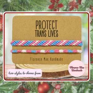 May include: Two woven bracelets featuring light blue, pink, and white threads displayed on a rectangular kraft paper card. The card displays the text PROTECT TRANS LIVES and Florence Mae Handmade. A small label reads two styles to choose from.