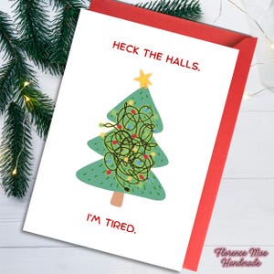 May include: A Christmas card with a red envelope features a green Christmas tree with tangled lights and a yellow star. The text reads "Heck the Halls. I'm Tired." The card is decorated with a sprig of evergreen and fairy lights.