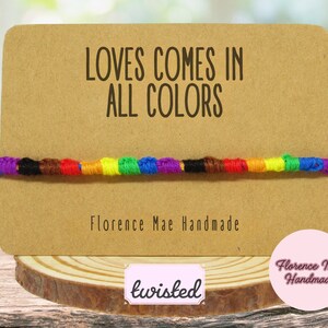 May include: A multi-colored knotted string bracelet displayed on a rectangular tan card. The card features the text "LOVES COMES IN ALL COLORS" and "Florence Mae Handmade" in black font. The bracelet consists of segments of blue, black, brown, red, orange, yellow, green, and purple thread.