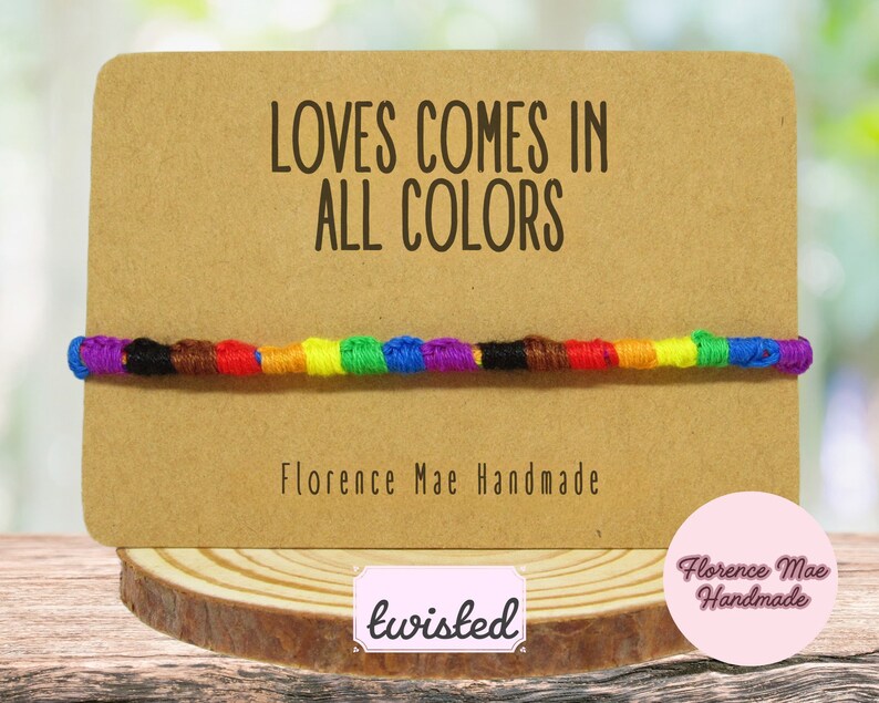 May include: A multi-colored knotted string bracelet displayed on a rectangular tan card. The card features the text "LOVES COMES IN ALL COLORS" and "Florence Mae Handmade" in black font. The bracelet consists of segments of blue, black, brown, red, orange, yellow, green, and purple thread.