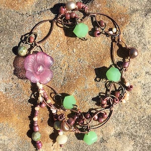 May include: A copper wire bracelet with a pink flower charm, green glass beads, and brown beads. The bracelet is on a brown, textured surface.