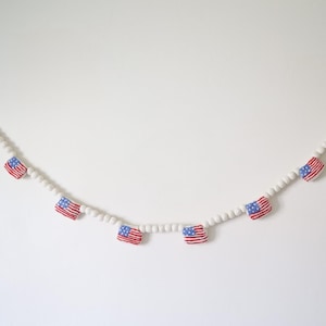 May include: A decorative garland featuring small American flags interspersed with white beads. The flags have red and white stripes with a blue square and white stars. The garland is strung on a thin, clear string.