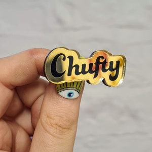 May include: Gold-colored enamel pin with the word "Chuffy" in black lettering. The pin has a shiny, reflective surface.