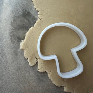 May include: A white plastic mushroom-shaped cookie cutter on a beige surface of rolled out cookie dough.