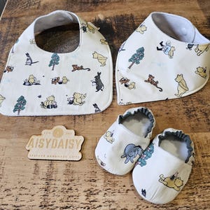 Puede incluir: Un juego de regalo para bebés que incluye un babero, un babero bandana y un par de zapatos para bebés. Todos los artículos son de color blanquecino y están decorados con personajes de dibujos animados y árboles. El letrero de madera dice "AisyDaisy Bespoke Handmade Gifts".