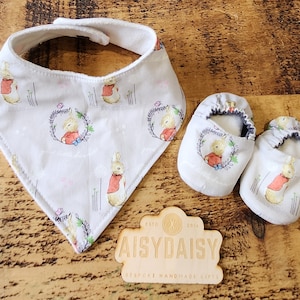 May include: A set of baby shoes and a bib with a white background and a repeating pattern of a rabbit in a red jacket. The shoes are gray and have a white trim. The bib is a triangle shape with a white trim. The set is made by AisyDaisy Bespoke Handmade Gifts.
