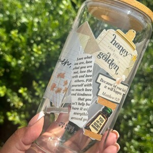 May include: A clear glass tumbler with a bamboo lid, decorated with various collage-style stickers featuring text such as honey your soul is golden, Matthew 9:29, and you are the secret ingredient, alongside illustrations of dried flowers and tape.