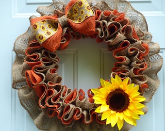 May include: A circular wreath constructed from ruffled tan burlap and orange fabric ribbons. The wreath features a large fabric bow at the top with a sunflower pattern and a single large, bright yellow artificial sunflower attached to the lower right.