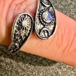 May include: A silver-toned ring with a celestial design. The ring features a sun and moon motif, with a textured surface and a blue gemstone. The ring is worn on a finger.
