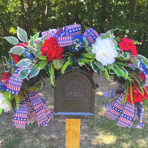 May include: A black mailbox with the text U.S. MAIL embossed on the front, adorned with a patriotic floral arrangement featuring red and white flowers, green foliage, and ribbons patterned with red, white, and blue stars and stripes.