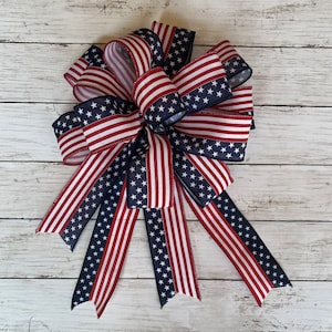 May include: A large red, white, and blue bow made of ribbon with a stars and stripes pattern. The bow is tied with a loop and tails.