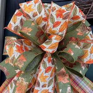 May include: A decorative bow made of wired ribbon featuring a pattern of autumn leaves in shades of orange, yellow, brown, and green. The design includes solid olive green ribbon sections and a multi-colored plaid ribbon at the bottom edges.