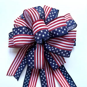 May include: A red, white, and blue bow with a stars and stripes pattern. The bow is made of fabric and has a wired edge. It is perfect for decorating for the Fourth of July or other patriotic holidays.
