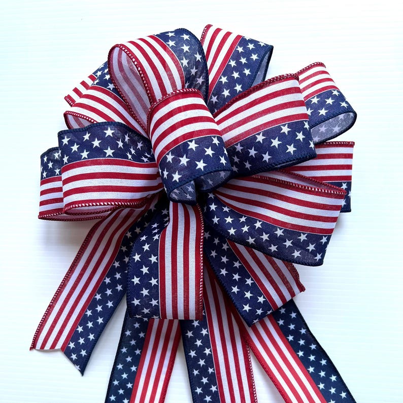 May include: A red, white, and blue bow with a stars and stripes pattern. The bow is made of fabric and has a wired edge. It is perfect for decorating for the Fourth of July or other patriotic holidays.