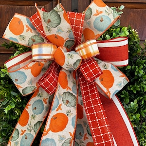 May include: A decorative bow featuring a fall harvest theme. The bow is made with ribbons in orange, red, and cream colors. The ribbons are patterned with pumpkins and plaid designs. The bow is attached to a green wreath.