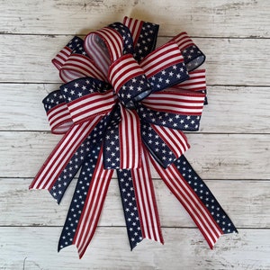 May include: A large red, white, and blue bow made of ribbon with a stars and stripes pattern. The bow is tied with a loop and tails.