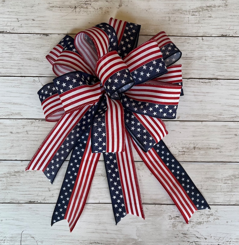 May include: A large red, white, and blue bow made of ribbon with a stars and stripes pattern. The bow is tied with a loop and tails.
