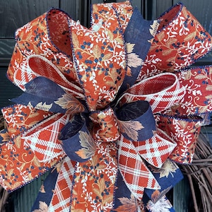 May include: A decorative bow featuring a mix of ribbons with orange, navy blue, and white colors. The patterns include a white floral and berry print on an orange background, a navy blue fabric with gold leaf accents, and a red and white plaid design.