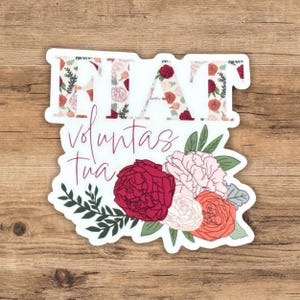May include: A white sticker with the words "Fiat Voluntas Tua" in pink script. The words "Fiat" are filled with a floral pattern in shades of red, pink, and white. A bouquet of flowers in shades of red, pink, and orange are below the text.