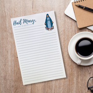 May include: White lined notepad with the text "Hail Mary, Full of Grace, The Lord is with Thee" in blue script. An illustration of the Virgin Mary in blue robes is at the top right, surrounded by flowers. The notepad is on a wooden surface.