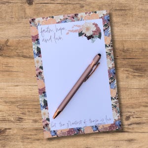 May include: A white notepad with a floral border and the quote "faith, hope, and love, but the greatest of these is love." (1 Corinthians 13:13) A rose gold pen is lying on the notepad.