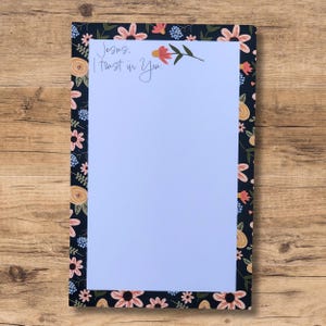 May include: A white notepad with a black floral border and the text "Jesus, I trust in You." The floral border features pink, yellow, and blue flowers on a black background.