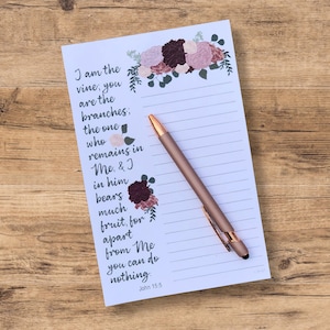 May include: A white notepad with a floral design and lined pages. The notepad features a quote from the Bible, John 15:5, "I am the vine, you are the branches; the one who remains in Me, & I in him bears much fruit, for apart from Me you can do nothing."