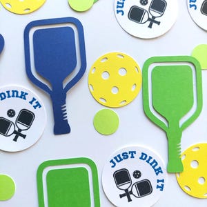May include: A set of party decorations for a pickleball themed party. The decorations include green and blue pickleball paddles, yellow pickleballs, and white circles with the text "Just Dink It" in blue.