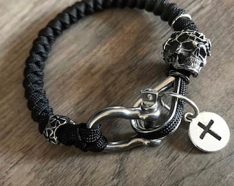 May include: A black paracord bracelet with a silver skull charm, a silver clasp, and a silver charm with a cross.