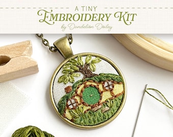 May include: A tiny embroidery kit by Dandelion Doiley. The kit includes a round pendant with an embroidered scene of a hobbit hole, a needle, and green thread. The pendant is on a chain. Additional items include a wooden clamp and embroidery hoop.