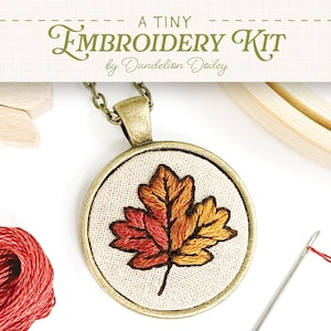 May include: A Tiny Embroidery Kit by Dandelion Doiley. The image shows a pendant with an embroidered maple leaf in shades of red, orange, and yellow. The pendant is on a chain, with a needle and thread, embroidery hoop, and red thread in the background.