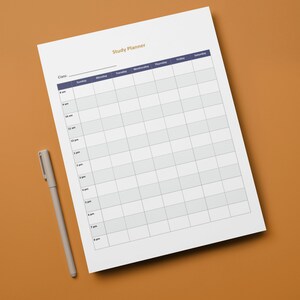 May include: A white study planner with a grid layout and the title "Study Planner" in gold lettering. A grey pen rests beside the planner on a brown surface. The planner has a dark blue header with the days of the week.