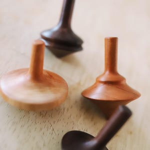 May include: Four wooden spinning tops, two are brown and two are light brown. The tops are on a light wood surface.