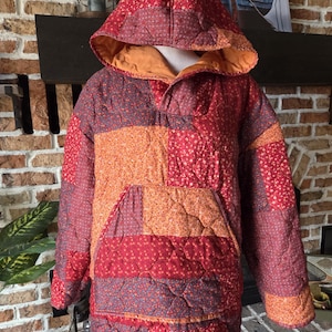 May include: A quilted pullover hoodie featuring a patchwork design with various floral and geometric patterns in shades of red, orange, and dark brown. The garment includes a large front kangaroo pocket, long sleeves, and a hood with an orange lining.