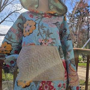May include: A quilted hoodie with a floral pattern in shades of blue, yellow, pink, and red. The hoodie features a large front pocket and a hood. The fabric appears to be soft and comfortable, suitable for casual wear.