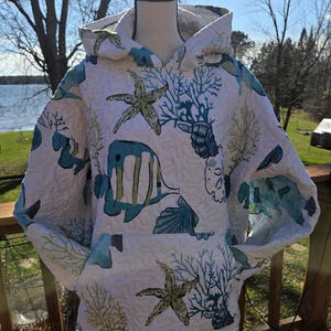 May include: White quilted hoodie with a blue and green sea life pattern, including starfish, fish, and coral. The hoodie has a front pocket and a hood. The background shows a lake and trees.