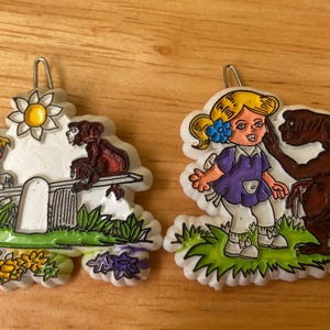 May include: Two white ceramic ornaments with colorful painted designs. One ornament features a girl in a purple dress with a flower in her hair, a brown creature with large ears, and green grass. The other ornament features a boy in a blue shirt, a girl in a red dress, a brown monkey on a seesaw, and a yellow flower.