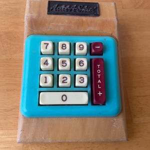 May include: A vintage blue and white toy calculator with a wooden frame. The calculator has a red button labeled "TOTAL +" and a black button labeled "-". The calculator has the words "Add A-Shiz" printed on the top.